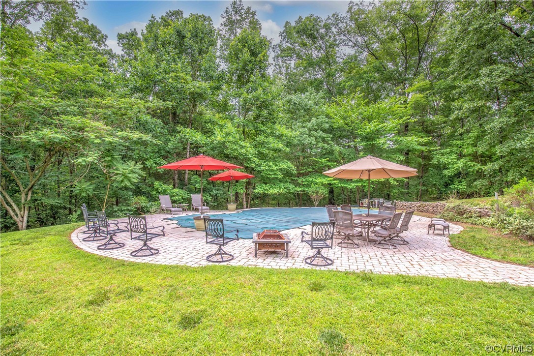 Backyard pool with a curved stone patio, outdoor seating, colorful umbrellas, and a wooded backdrop.
