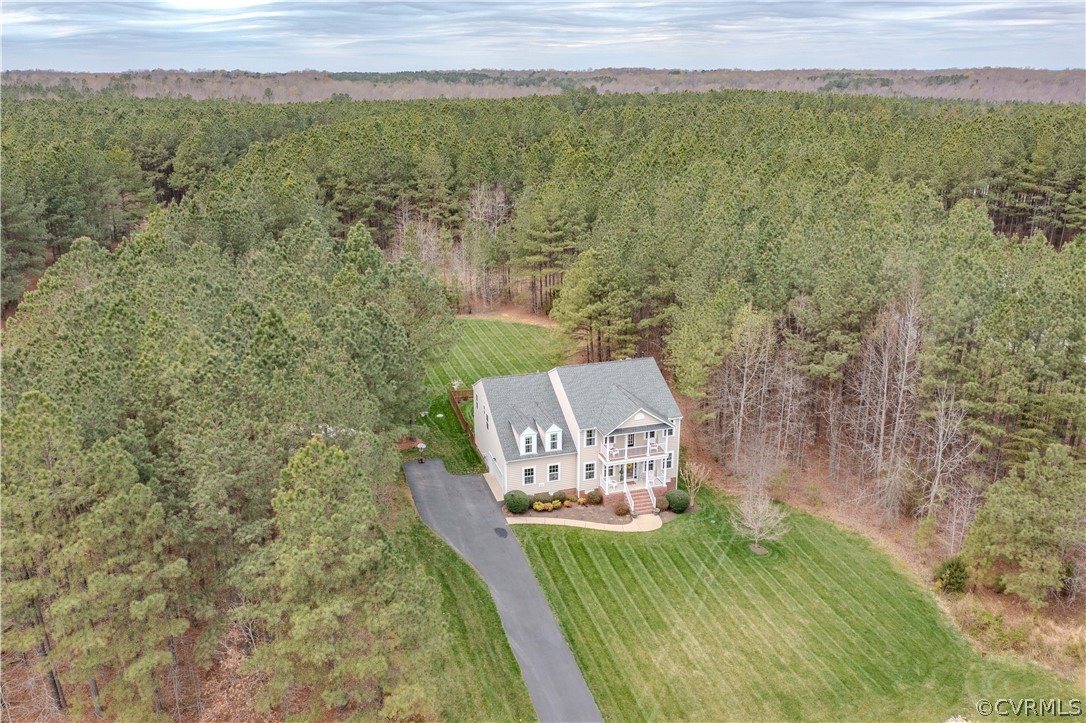 Aerial view of a two-story home with a striped lawn, long driveway, and surrounding woods.