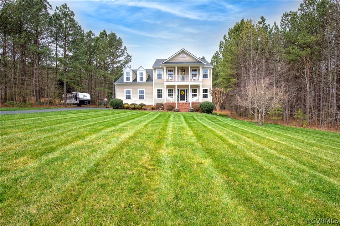Two-story beige house with an upper balcony, broad striped lawn, and wooded surroundings.