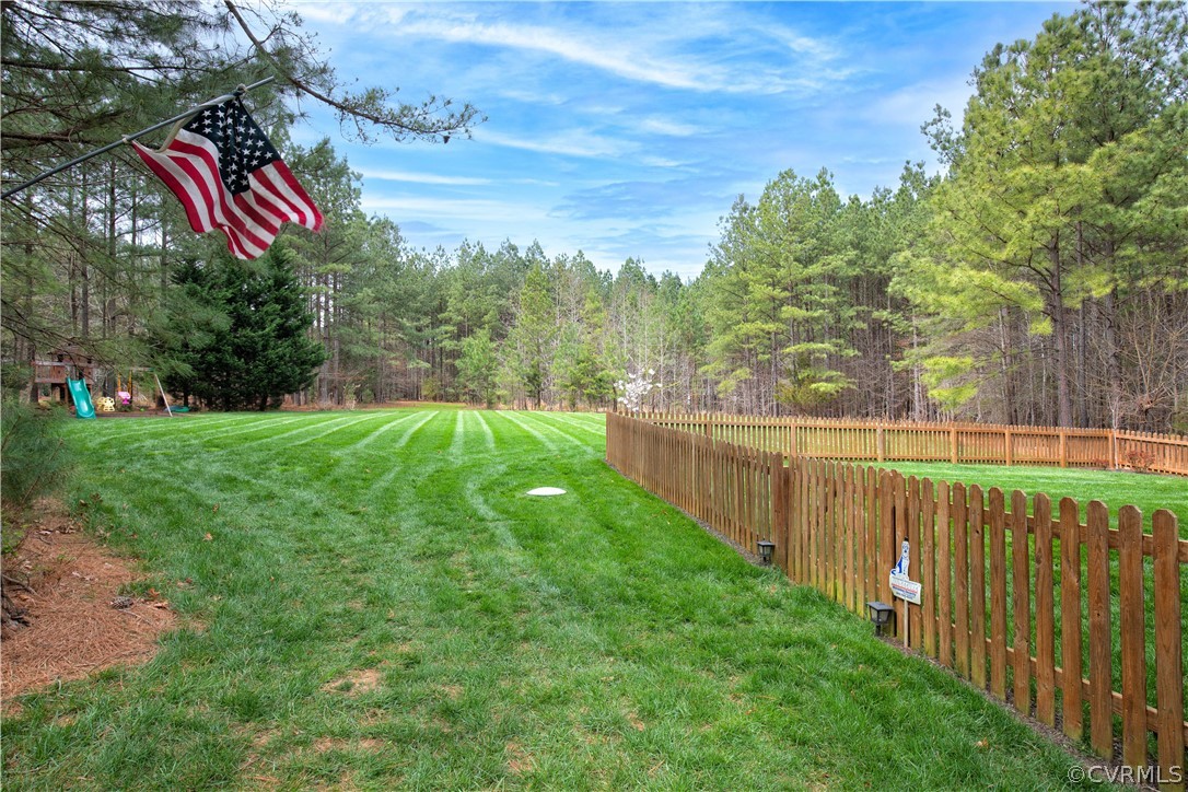 Fenced backyard with a striped lawn, wooded perimeter, and American flag.