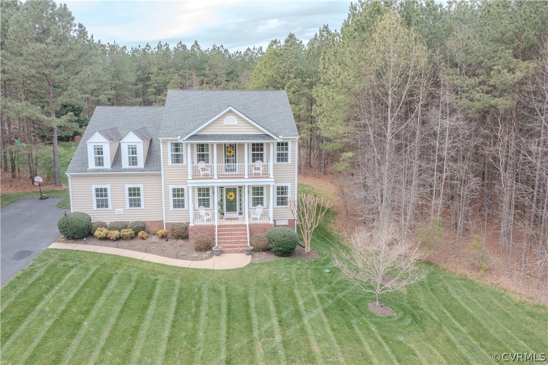 Aerial view of a beige two-story house with covered porches, a striped lawn, and wooded surroundings.