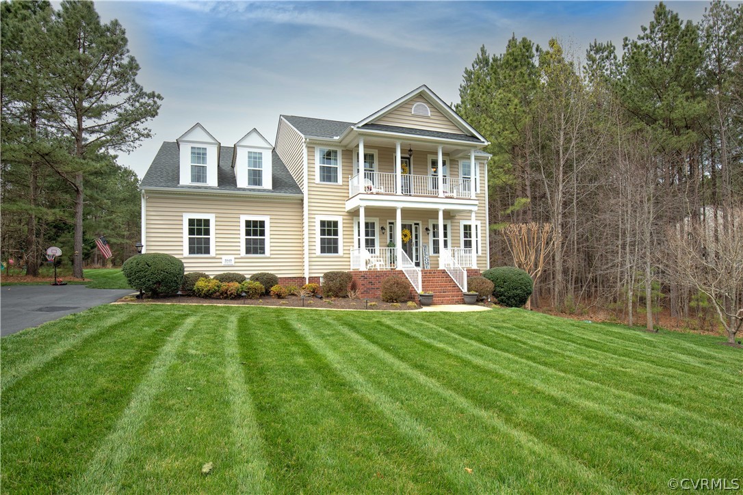 Two-story beige house with a double porch, dormer windows, and a broad striped lawn.