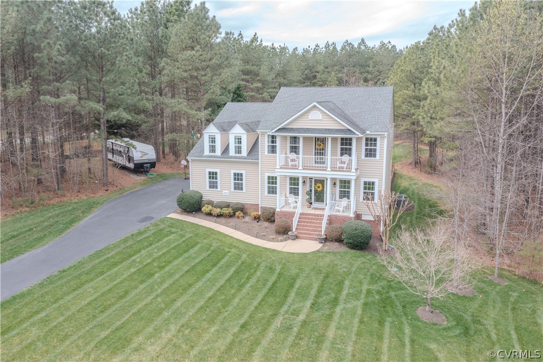 Two-story beige home with a front porch and balcony, striped lawn, driveway, and wooded surroundings.