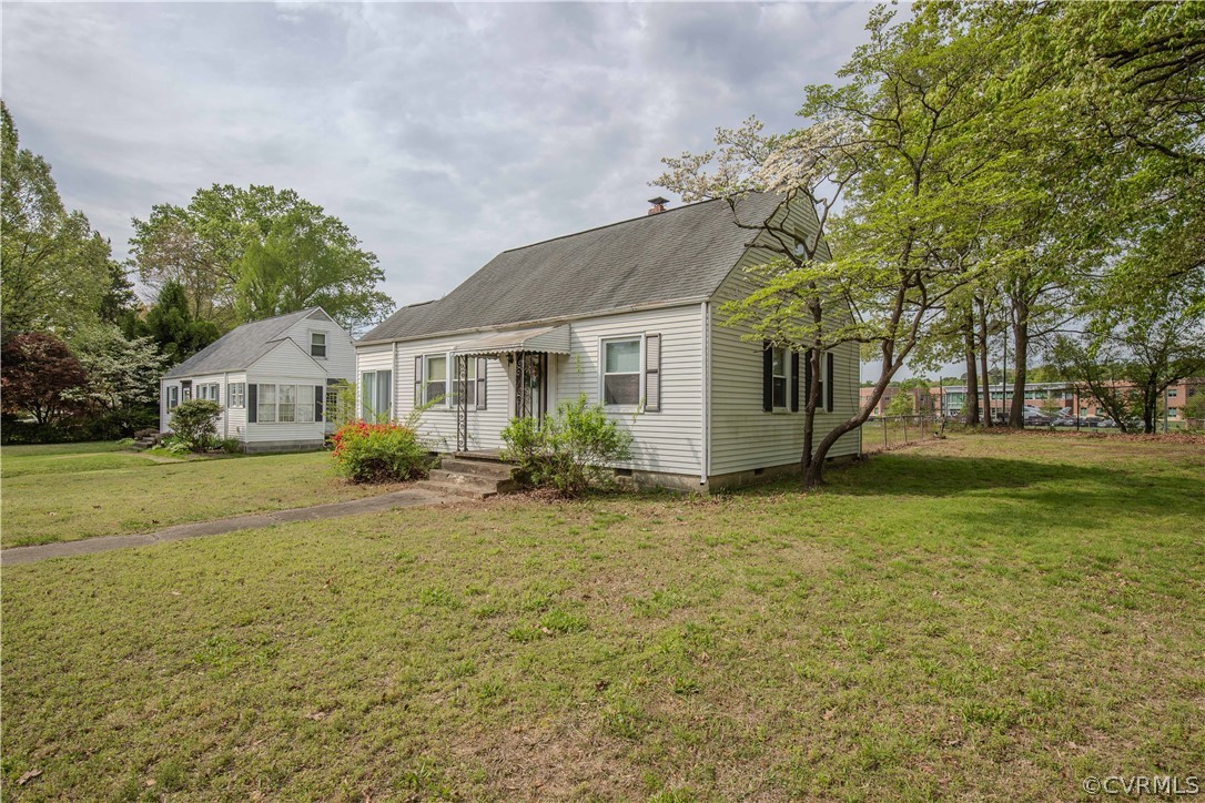 Single-story light-colored house with a gray roof, small outbuilding, and mature trees on a grassy lot.