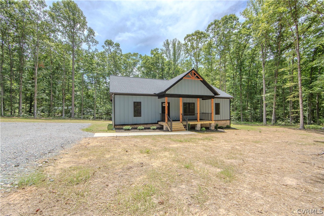 Gray-sided house with a covered porch, timber accents, and a gravel driveway in a wooded setting.