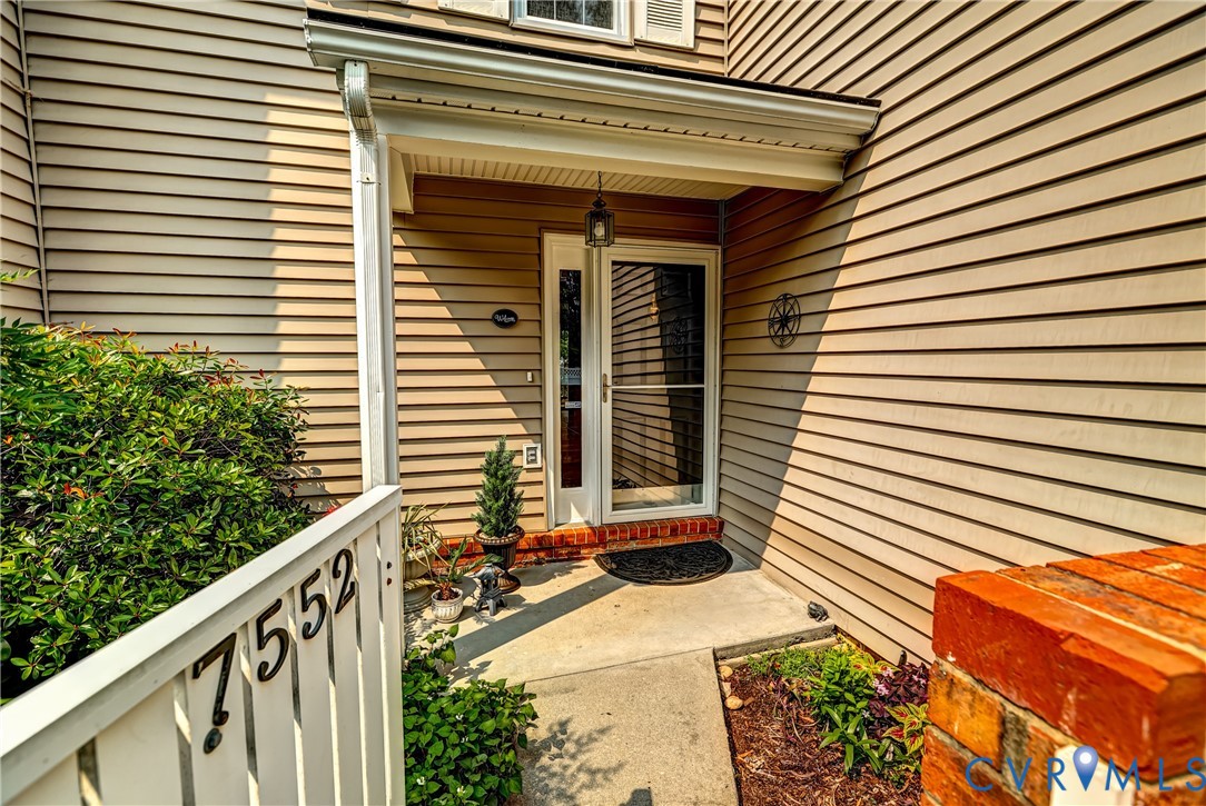 Covered front entry with tan siding, glass storm door, brick steps, and house number 7552.