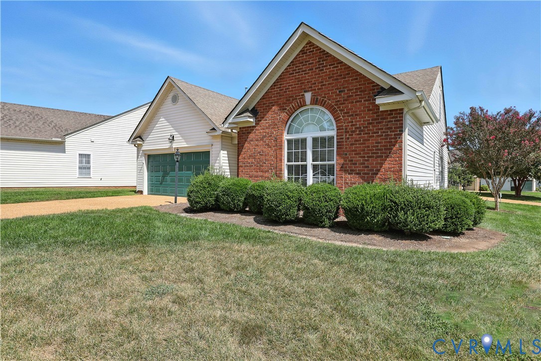 Brick-and-white-sided home with an arched front window, green double garage door, and trimmed shrubs.