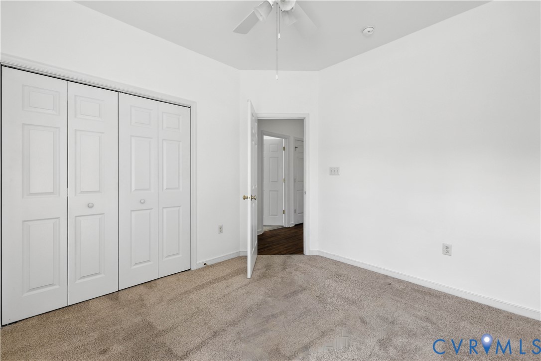 Empty bedroom with beige carpet, paneled closet doors, and an open doorway to a dark wood-floored hall.