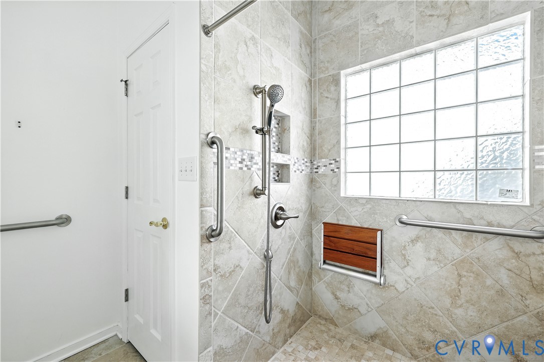 Tiled accessible shower with grab bars, handheld showerhead, fold-down seat, and frosted window.