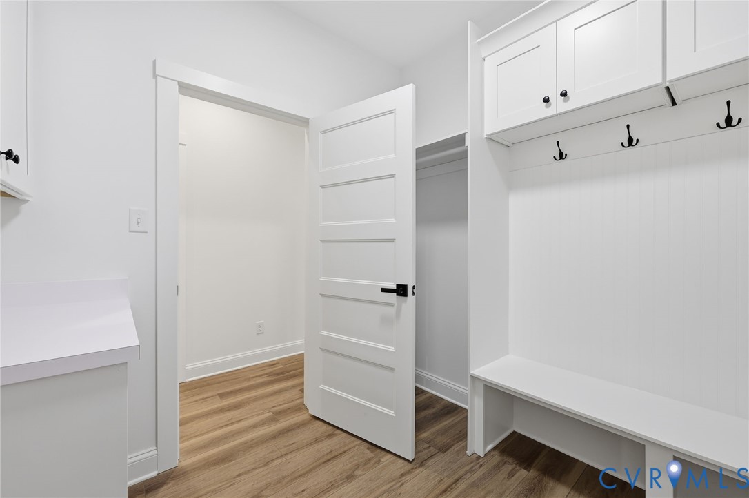 White mudroom with built-in bench, black coat hooks, overhead cabinets, and wood-look flooring.