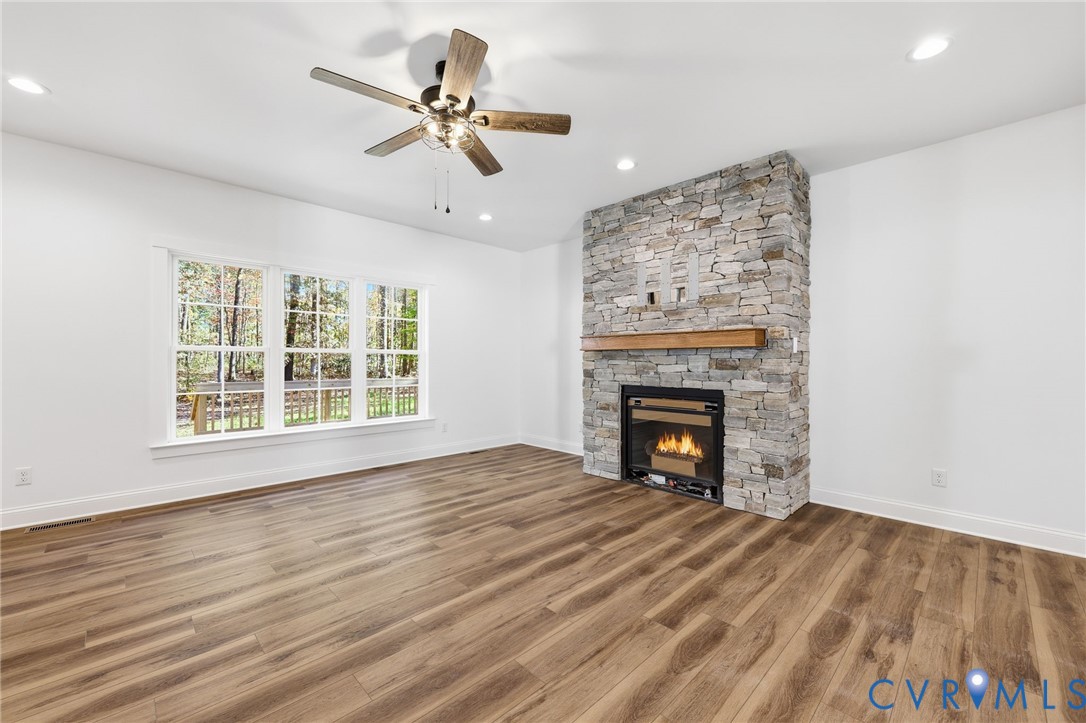 Vacant living room with a stone fireplace, wood floors, large windows, and ceiling fan.
