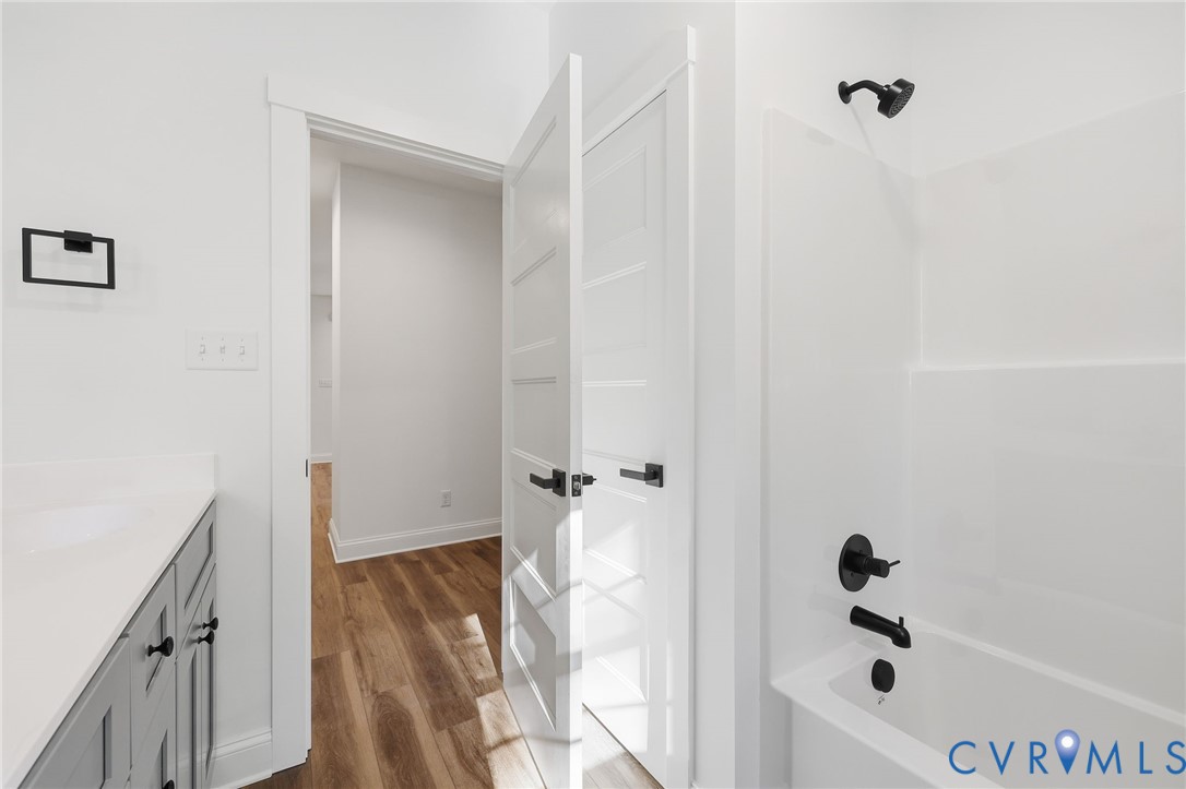 Bathroom with a white tub, black fixtures, gray vanity, and open door to a wood-floored hallway.