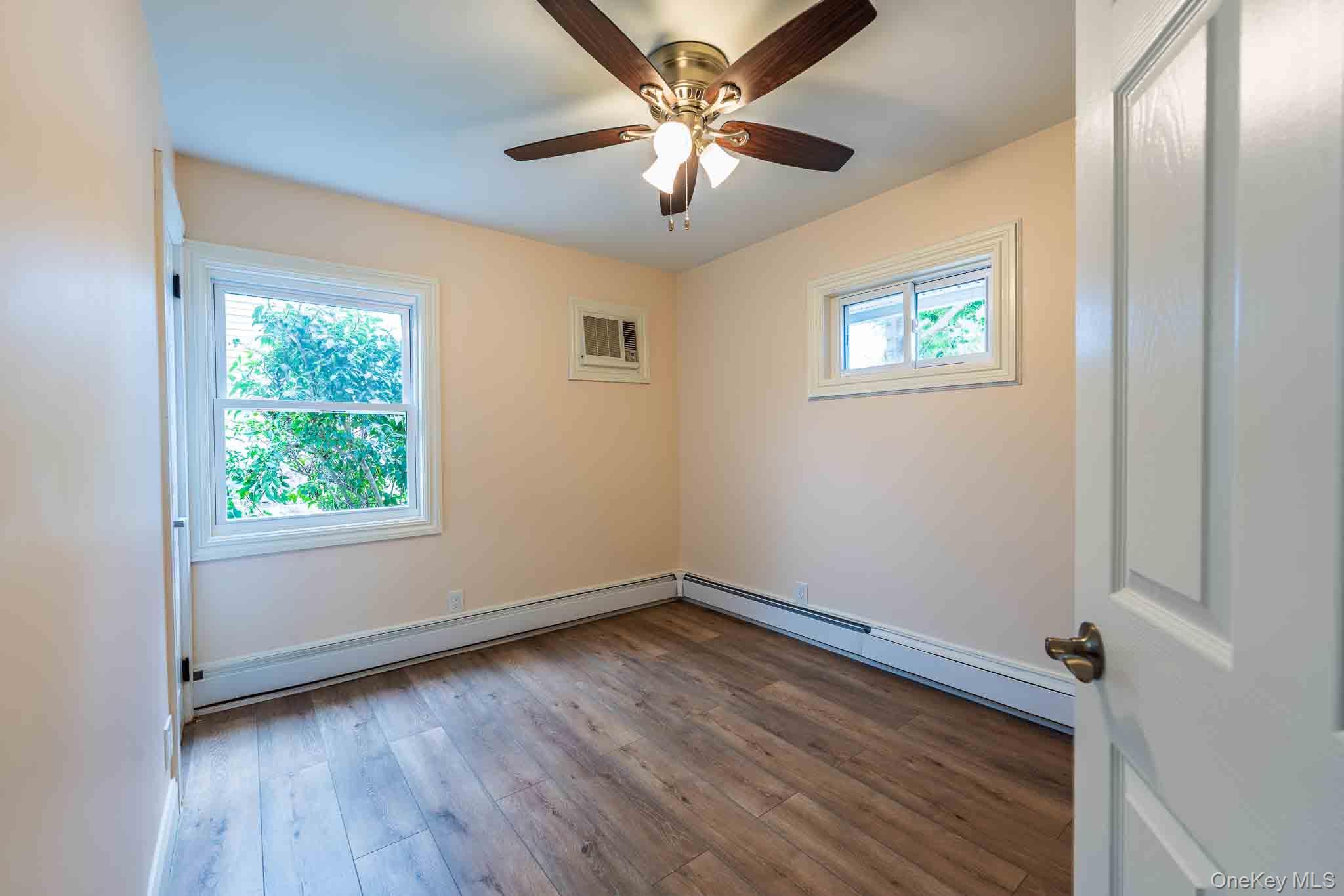Empty room with wood-look flooring, two windows, baseboard heating, and a ceiling fan.
