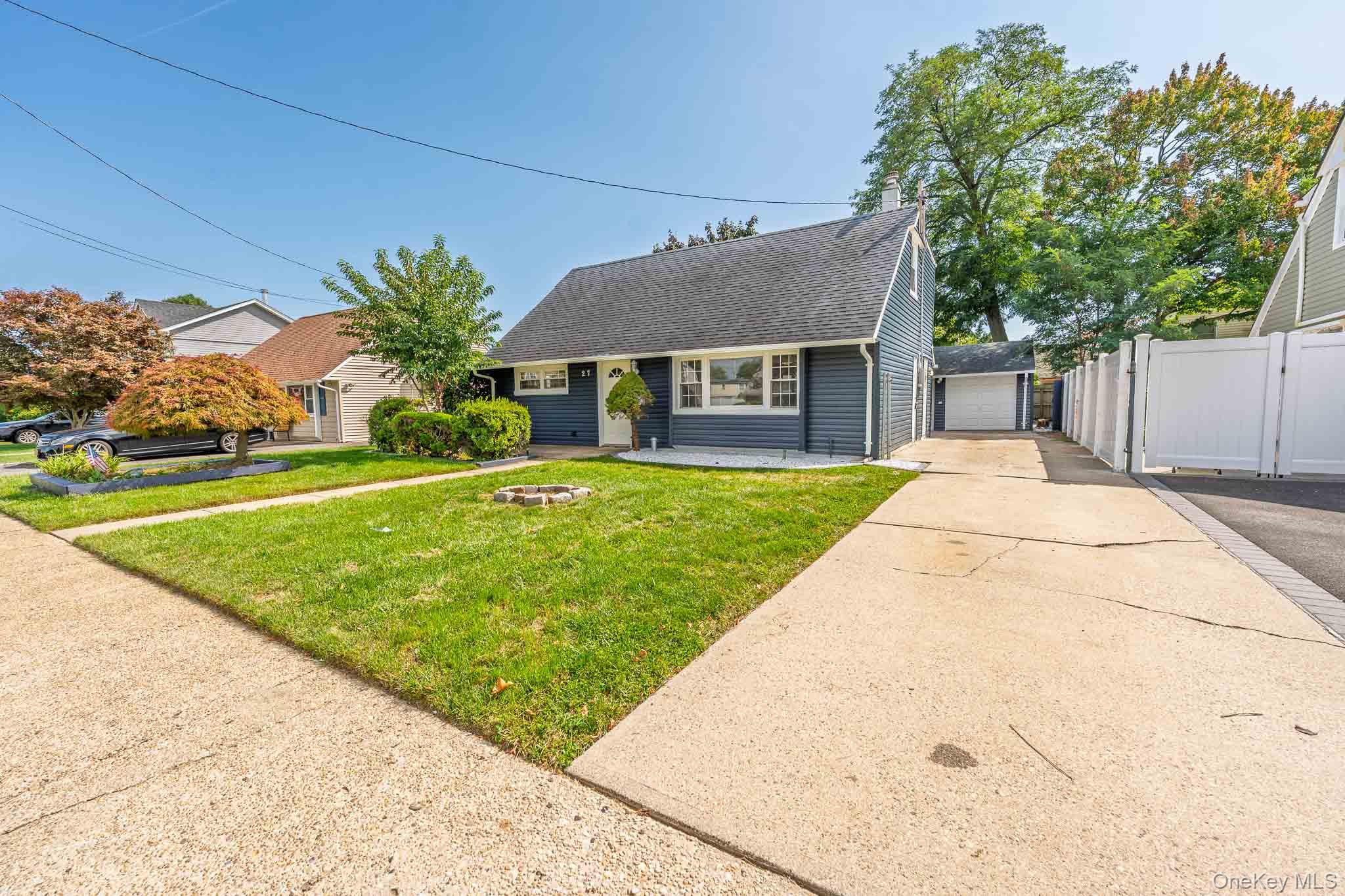 Dark blue Cape-style house with a green front lawn, long driveway, detached garage, and white privacy fence.