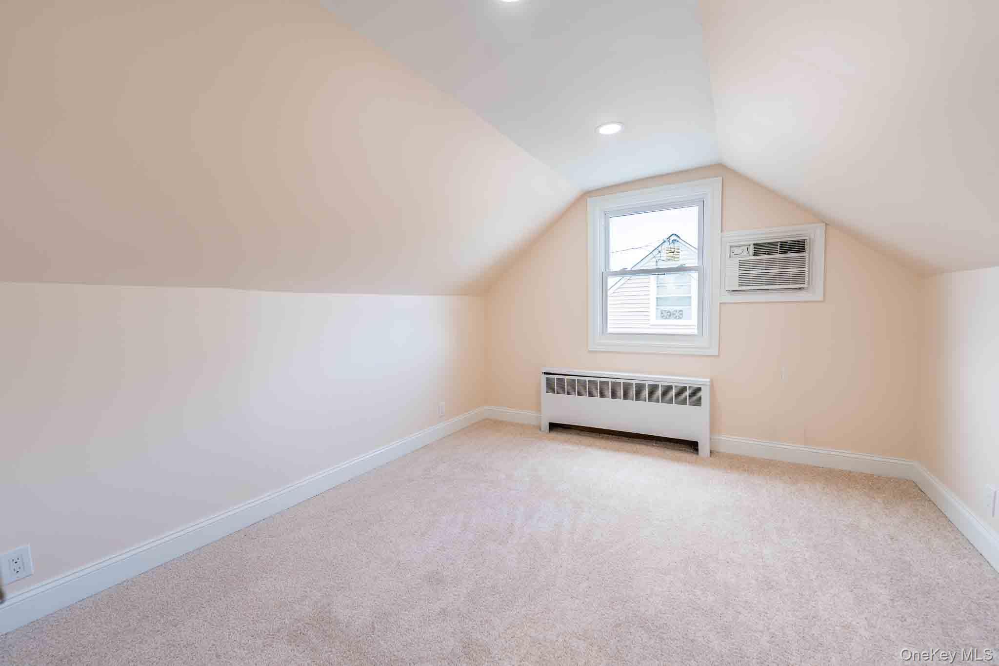 Empty carpeted room with a sloped ceiling, small window, and wall-mounted air conditioner.