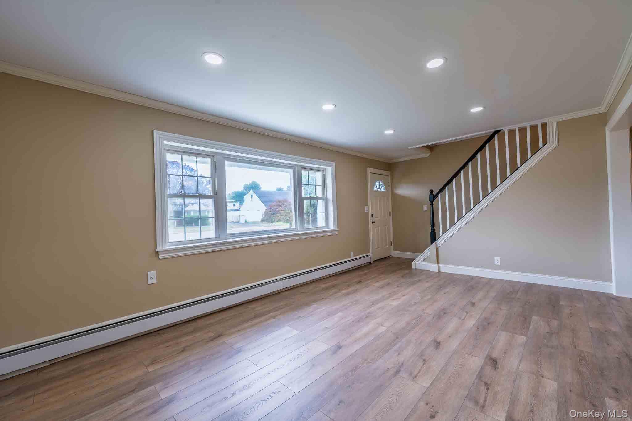 Empty living room with light wood floors, a large picture window, and a staircase beside the front door.