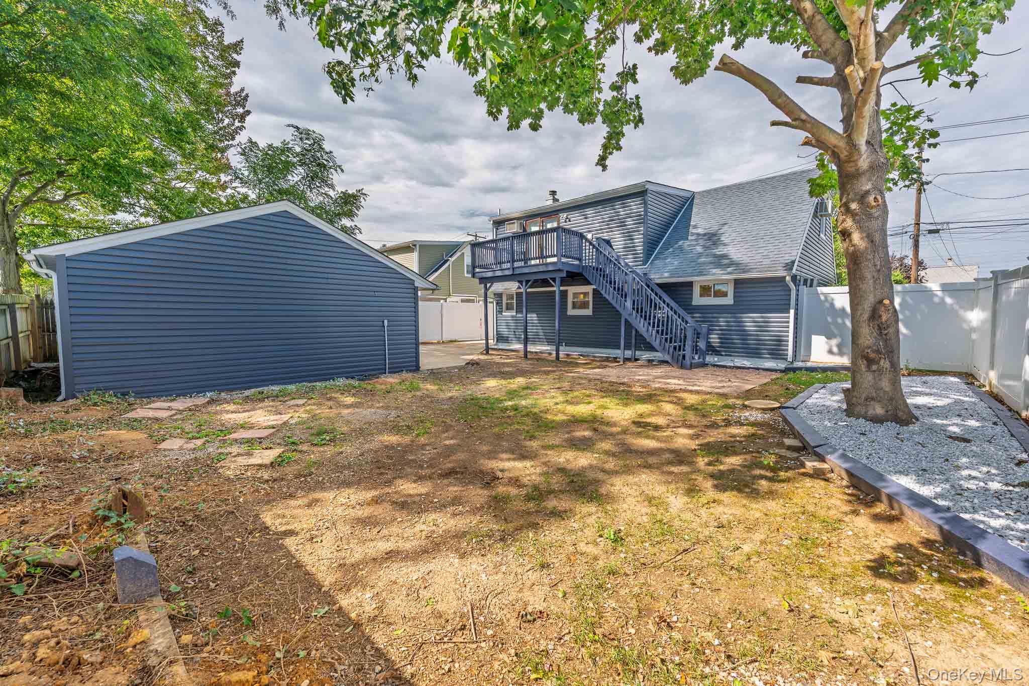 Blue house with a raised deck and staircase, detached garage, and fenced yard with a large tree.