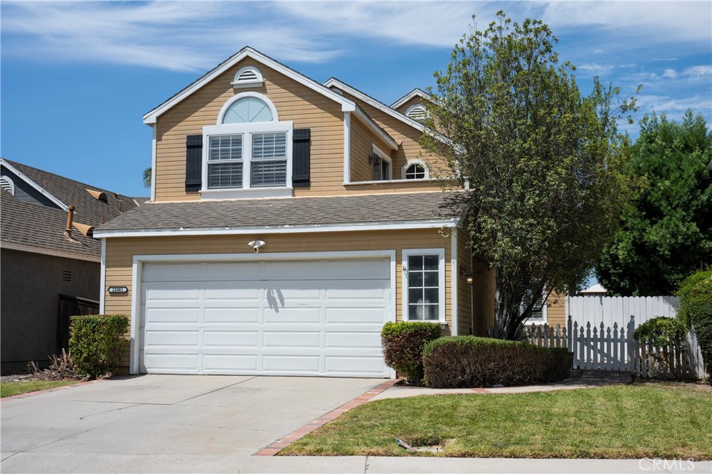 Two-story tan house with a white double garage, front lawn, and picket fence.