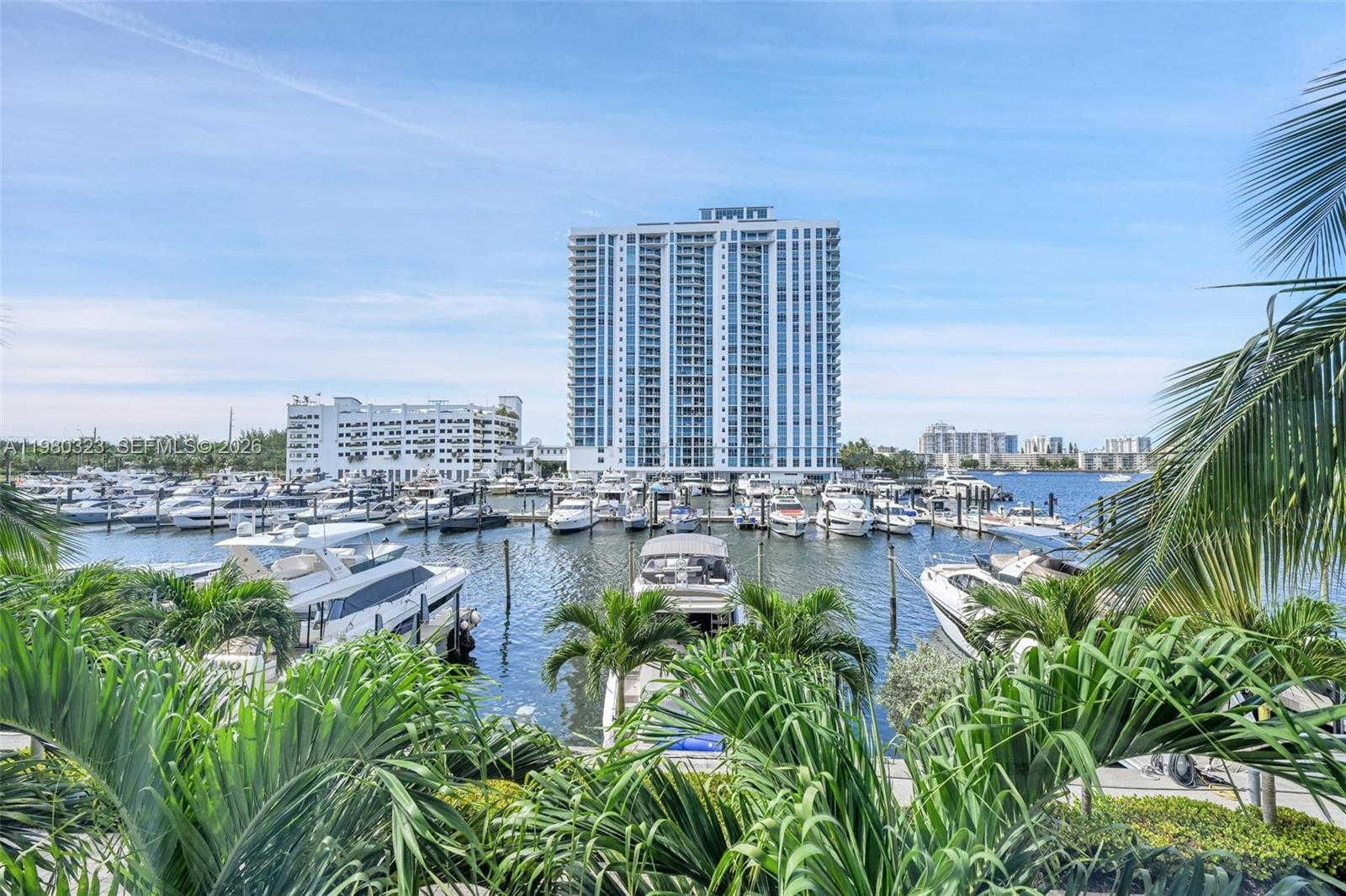 Waterfront condominium tower overlooking a marina filled with boats, framed by tropical palms.