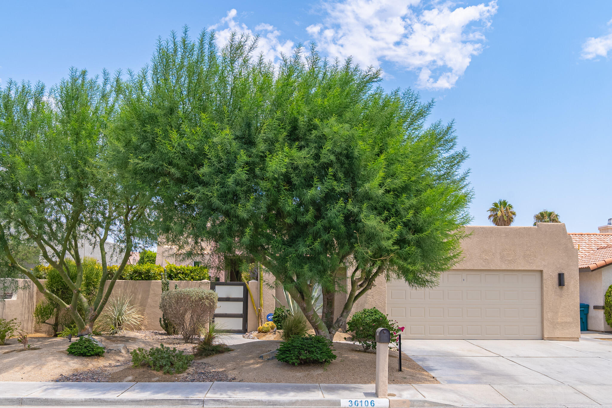 Stucco home with a double garage partly screened by large trees and desert landscaping.
