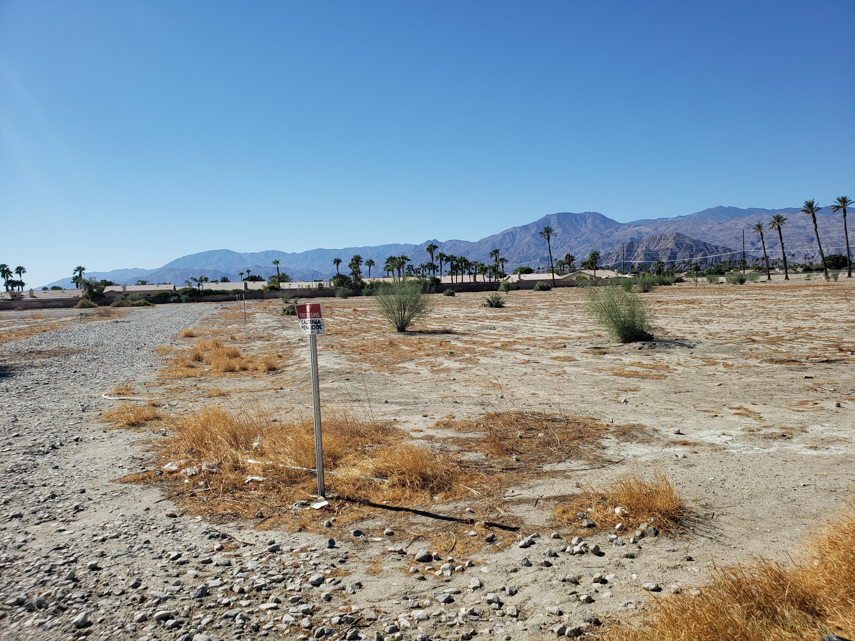 Vacant desert lot with a trespassing sign and mountains beyond palm-lined homes.