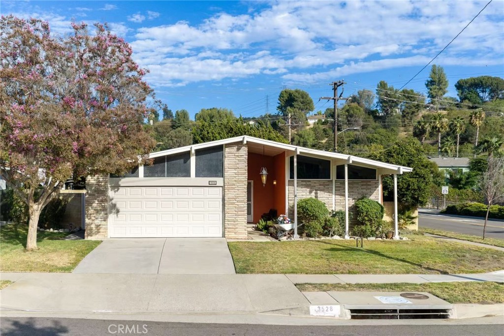 Single-story mid-century home with stone facade, broad garage, covered entry, and flowering tree.