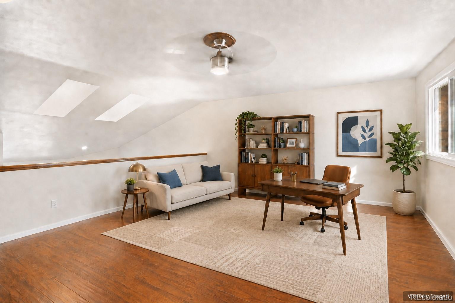 Staged upstairs home office with skylights, a wooden desk, bookcase, and cream sofa.