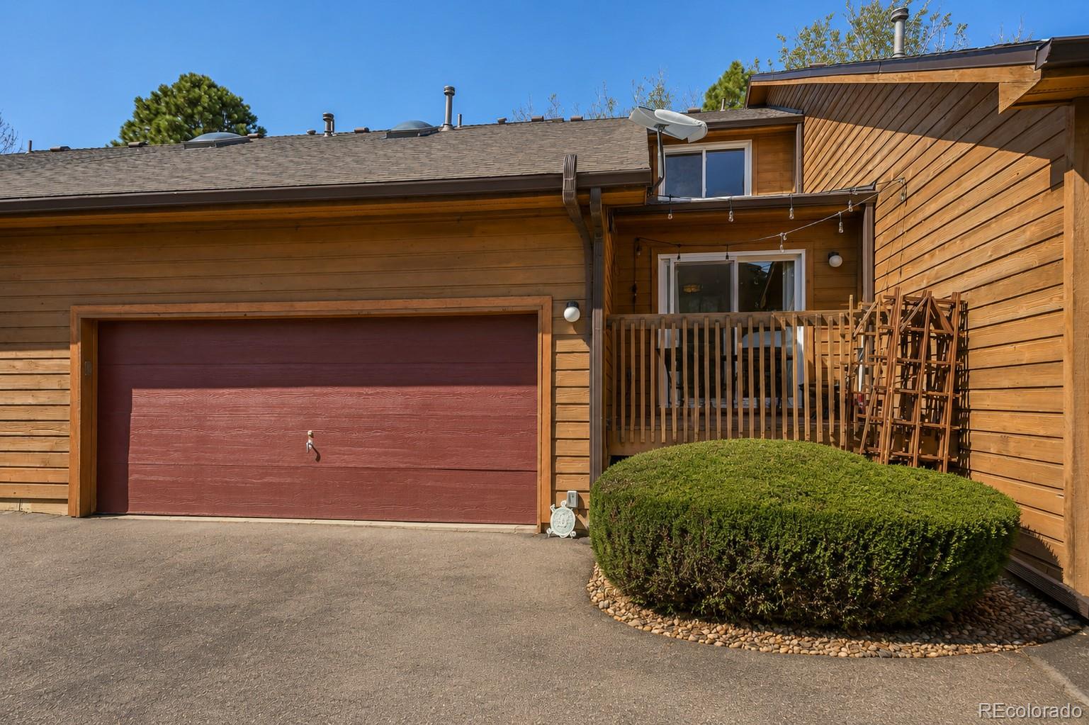 Wood-sided home exterior with burgundy garage door, raised deck, and rounded evergreen shrub.