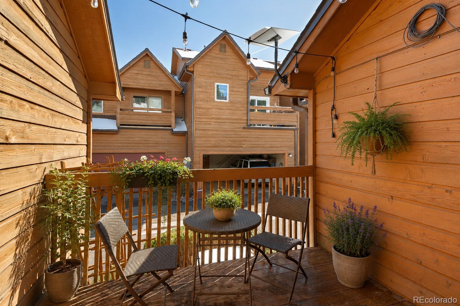 Wooden balcony with bistro seating, potted plants, string lights, and views of neighboring townhomes.