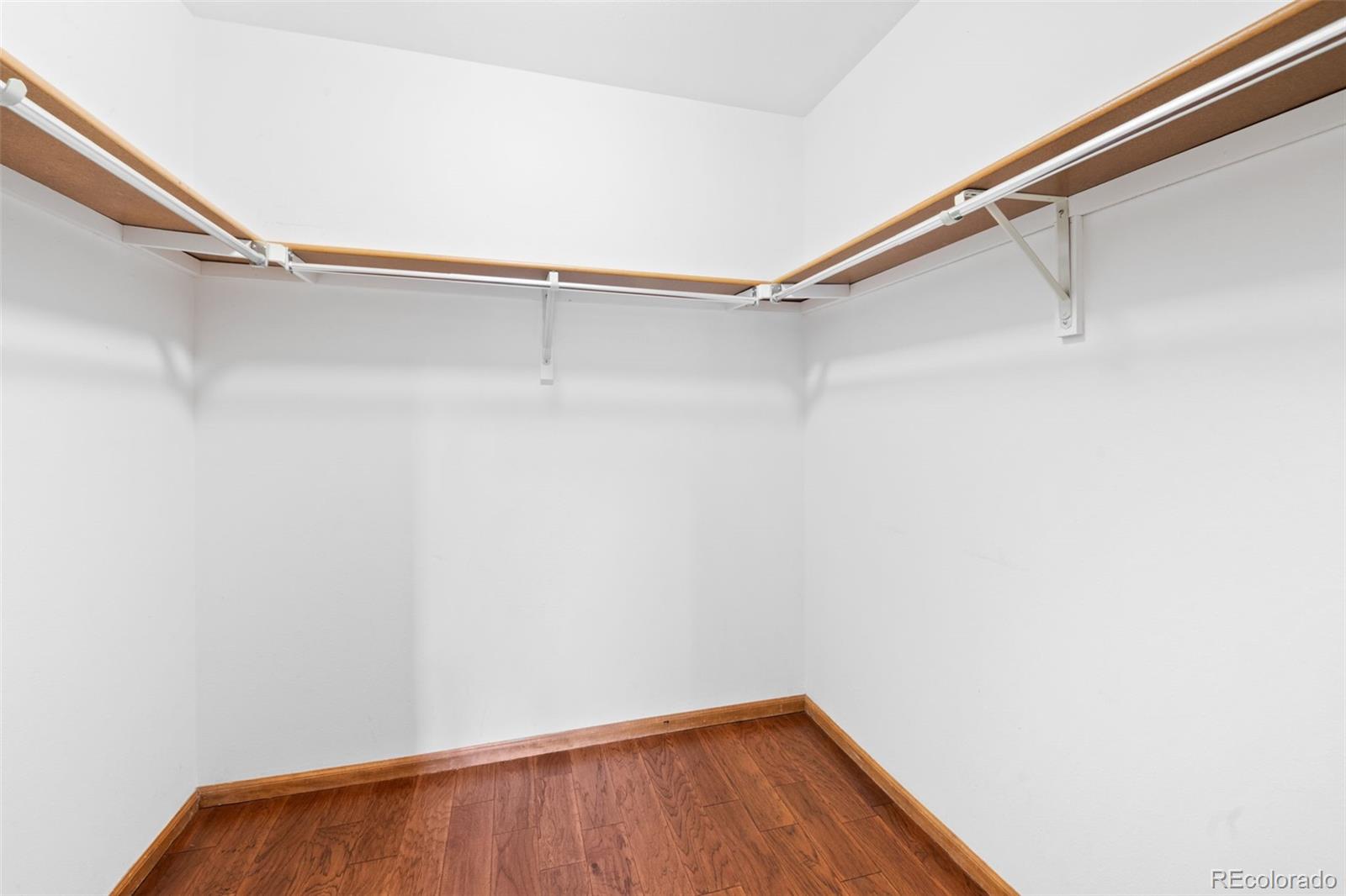 Empty walk-in closet with wraparound shelves and hanging rods above hardwood flooring.
