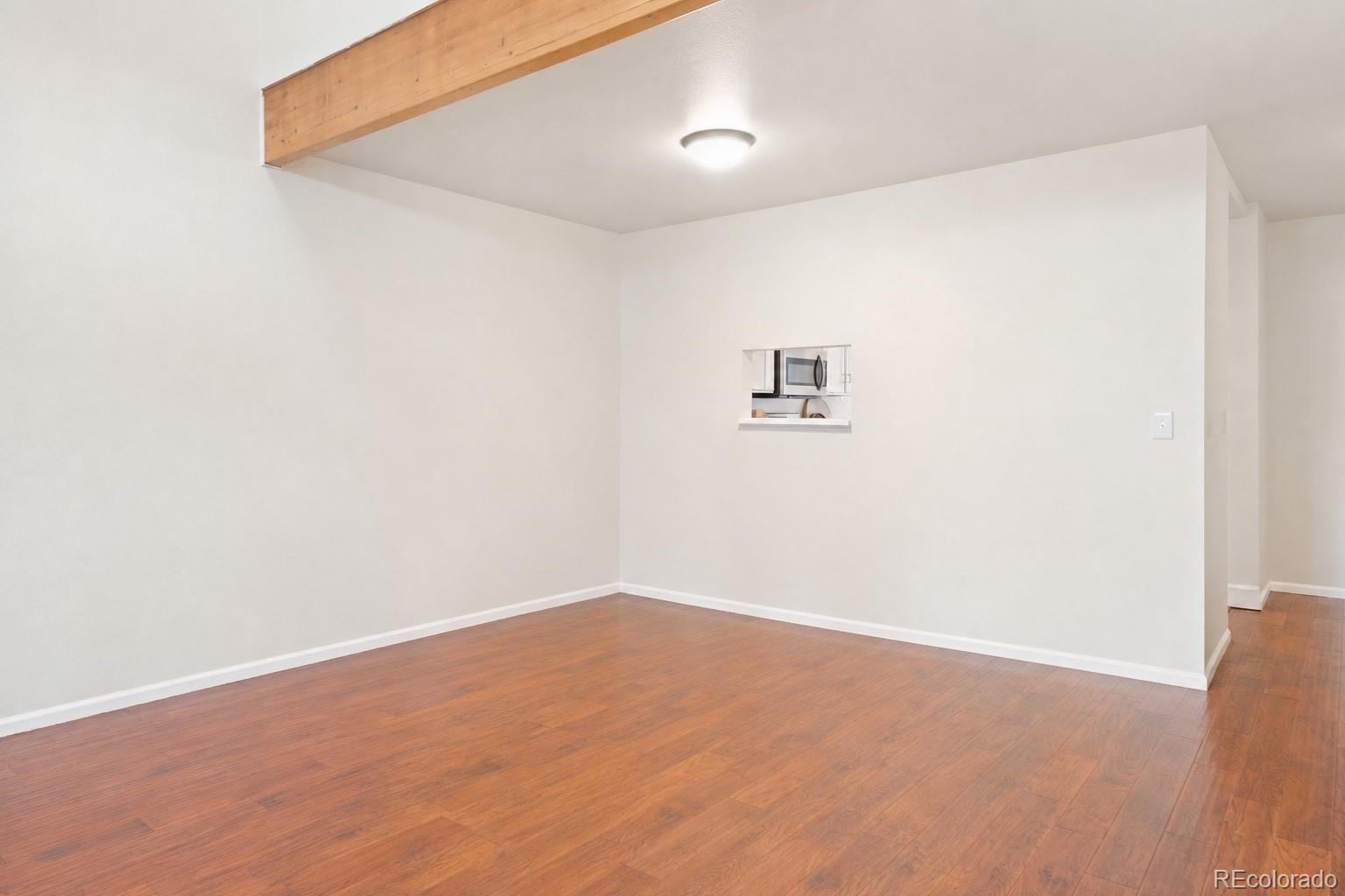 Empty living area with wood floors, an exposed ceiling beam, and a kitchen pass-through.