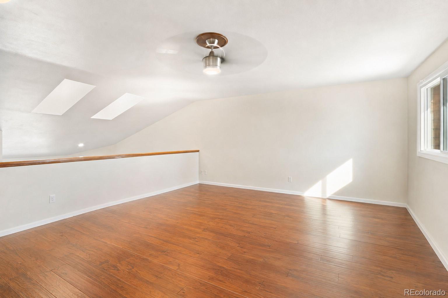 Empty loft with hardwood floors, two skylights, a sloped ceiling, and a ceiling fan.