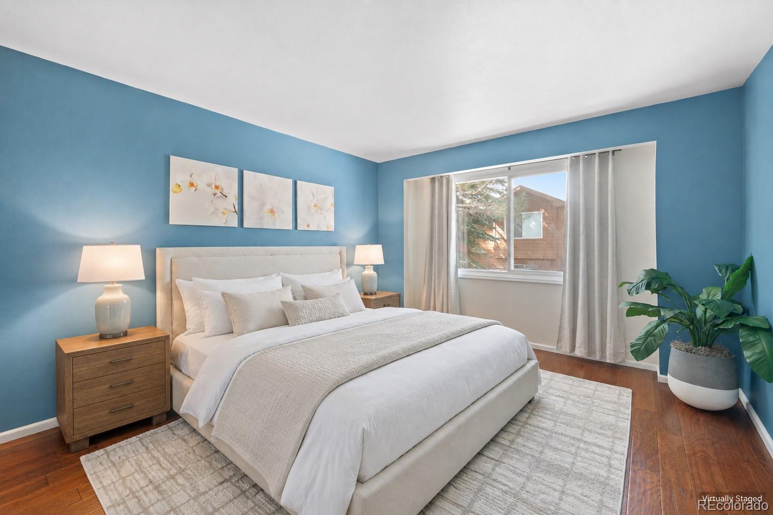 Staged bedroom with a white bed, blue walls, floral art, and a large curtained window.