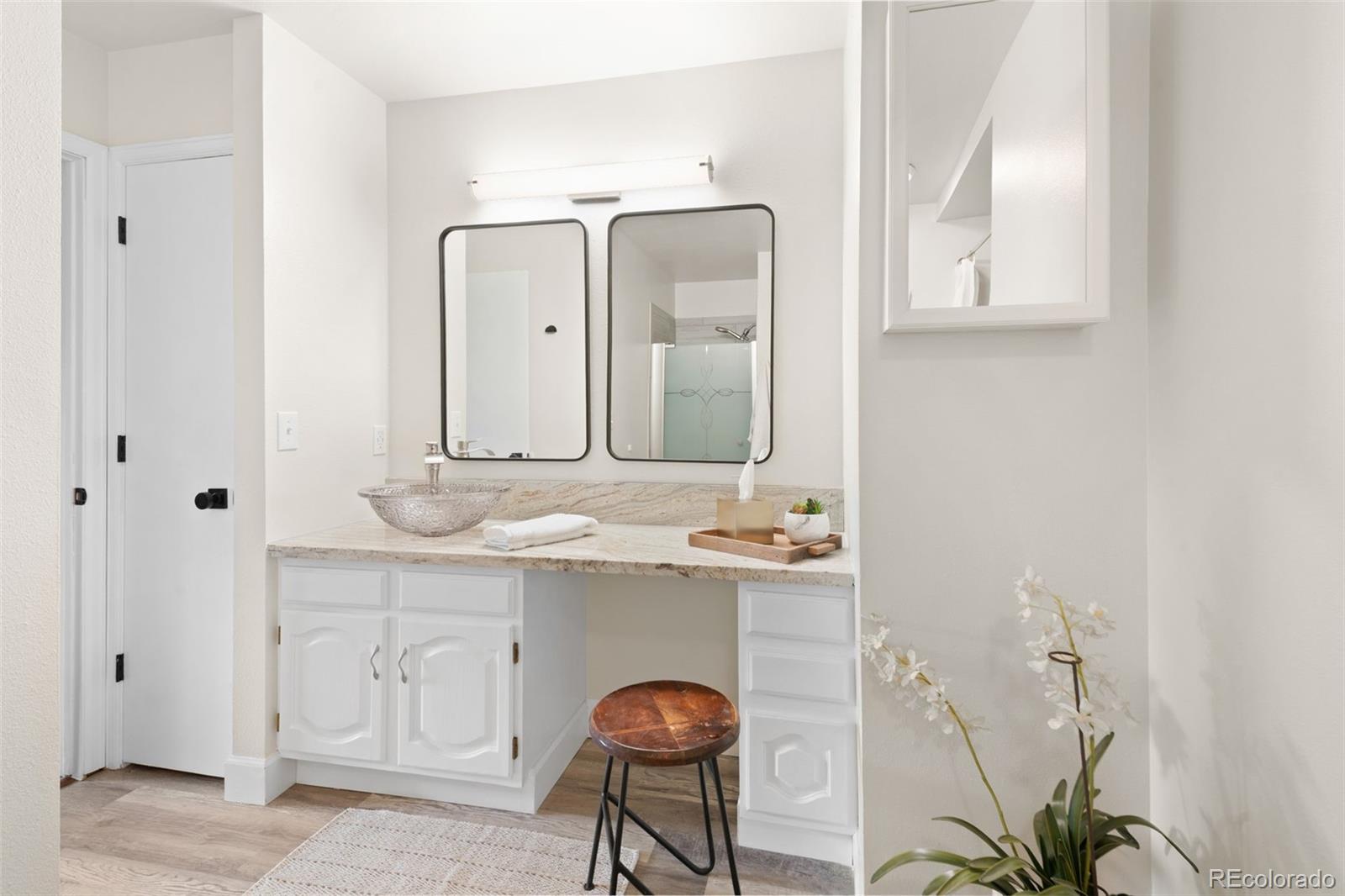Bathroom vanity with a glass vessel sink, stone counter, black-framed mirrors, and stool.