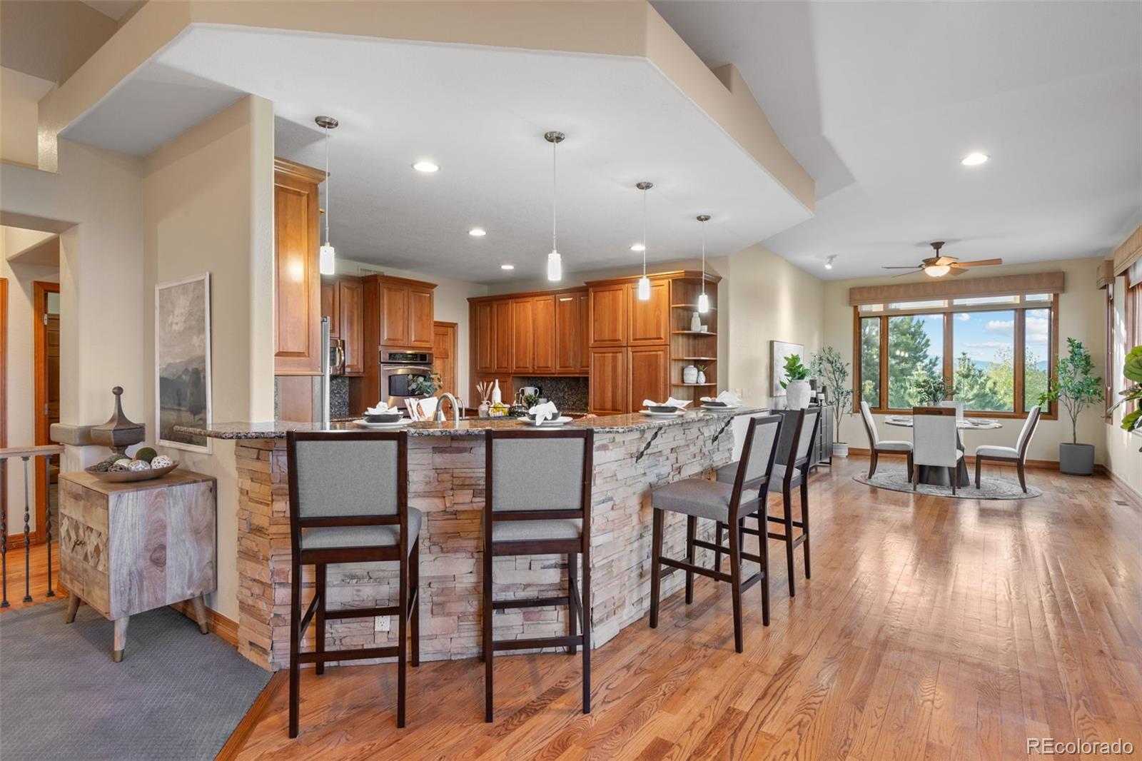 Open-plan kitchen and dining area with a stone island, warm wood cabinets, and mountain views through broad windows.