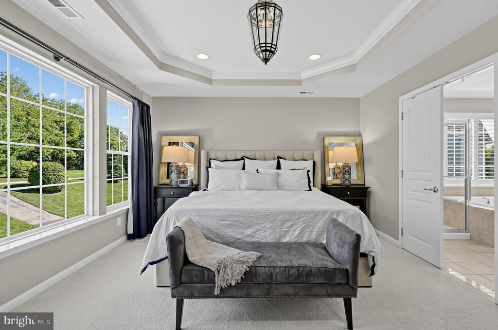 Staged bedroom with a large bed, gray bench, tray ceiling, and tall windows overlooking the lawn.