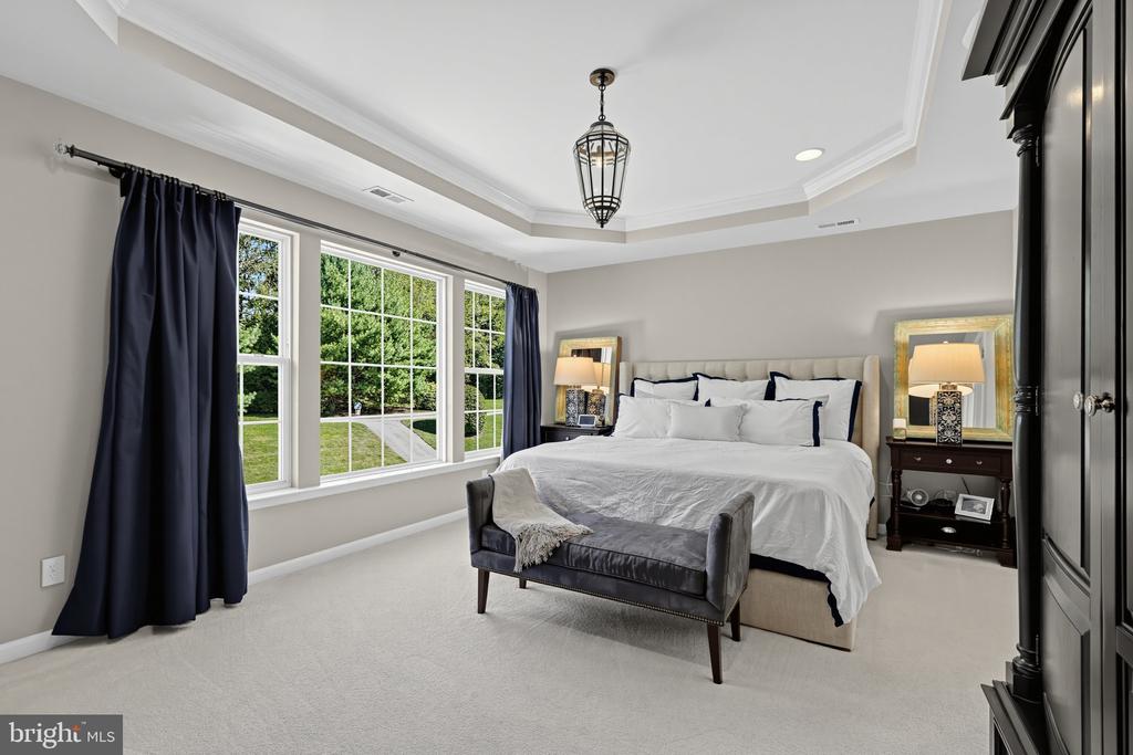 Primary bedroom with a tray ceiling, wide windows, dark blue curtains, and a gray upholstered bench.