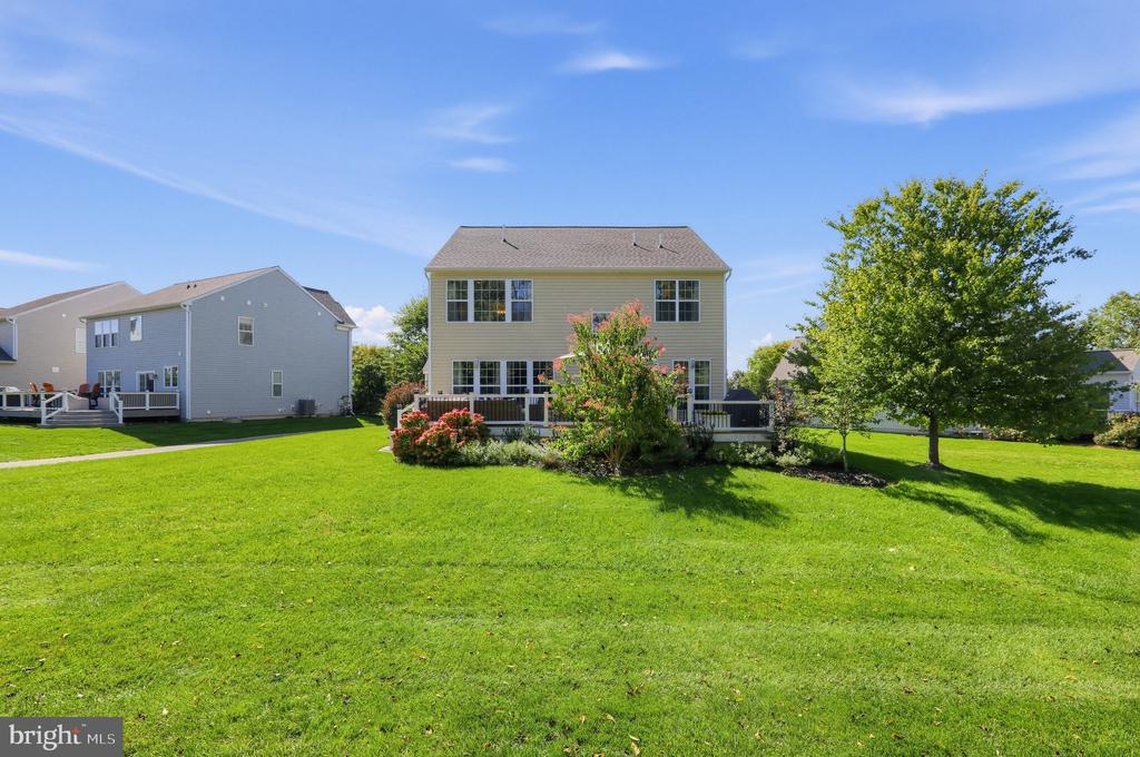 Two-story gray house with a raised deck, flowering shrubs, and a broad green lawn.