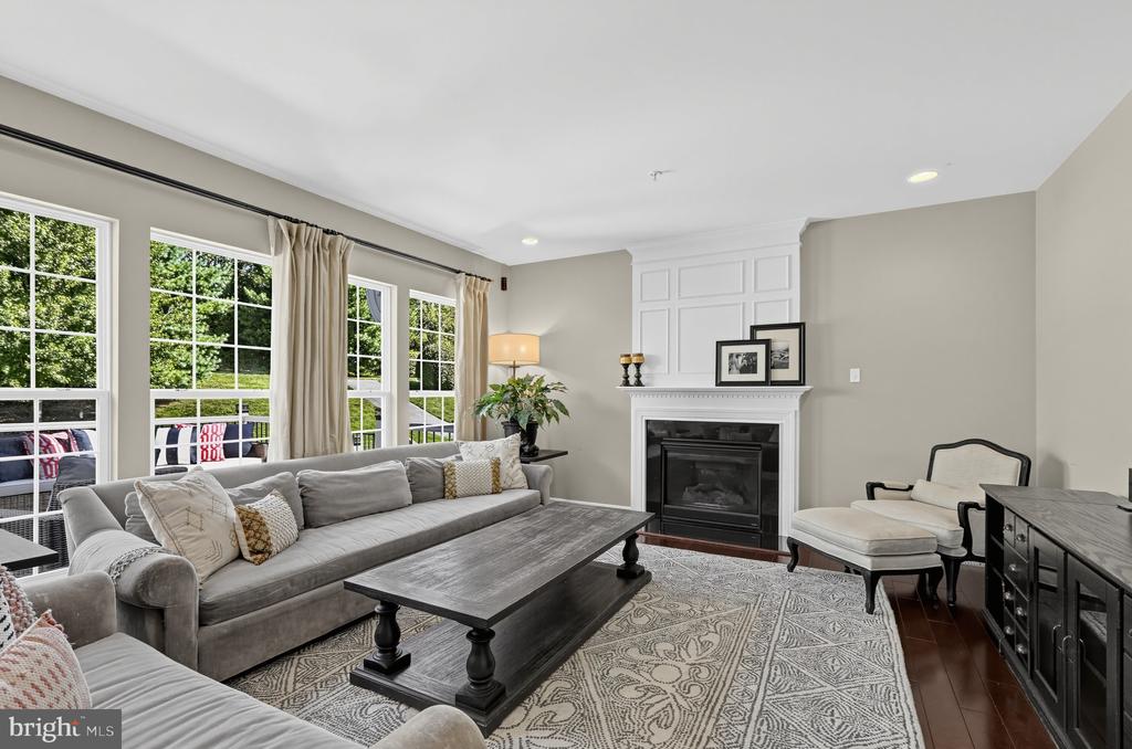 Living room with a gray sectional, white fireplace, and tall windows overlooking greenery.
