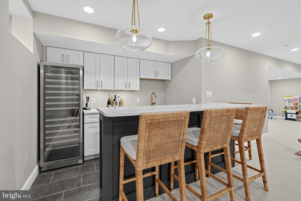 Modern kitchen with a dark island, woven bar stools, and a glass-front wine refrigerator.