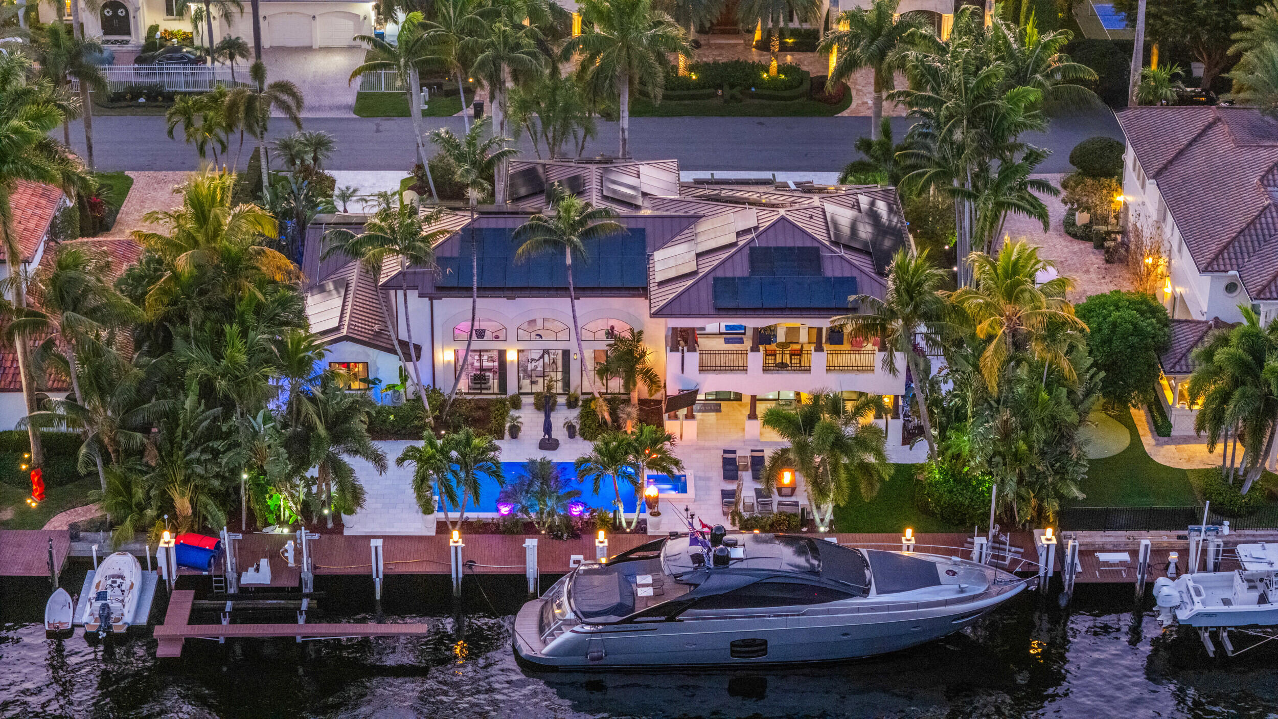 Aerial view of a waterfront home with a lit pool, palm trees, and a yacht at the dock.