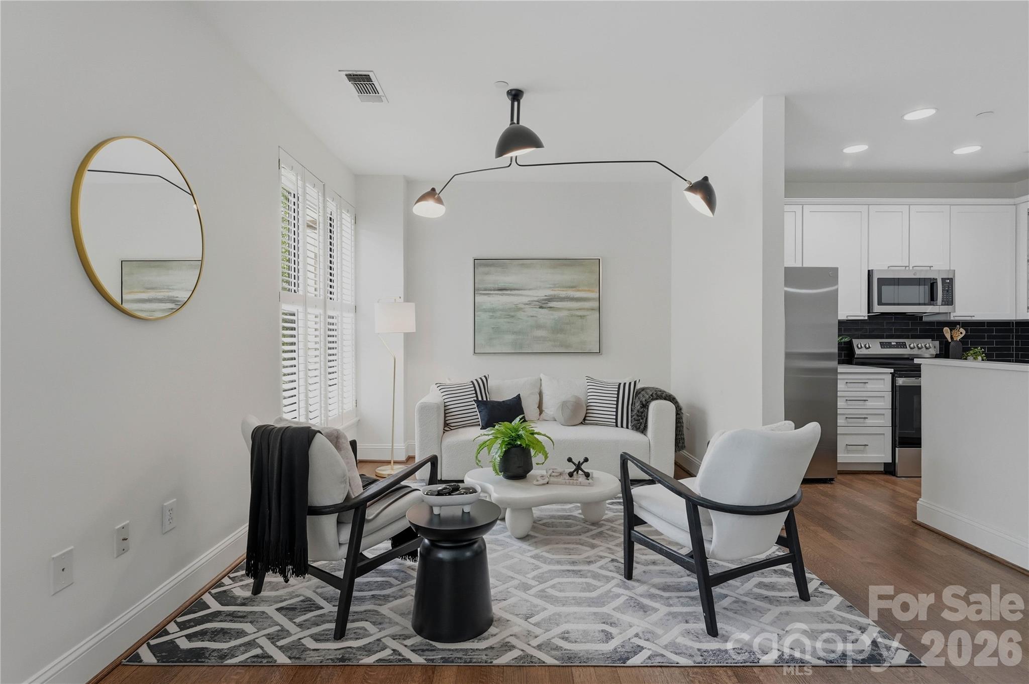 Furnished living room opening to a white kitchen, with a geometric rug and gold-framed mirror.