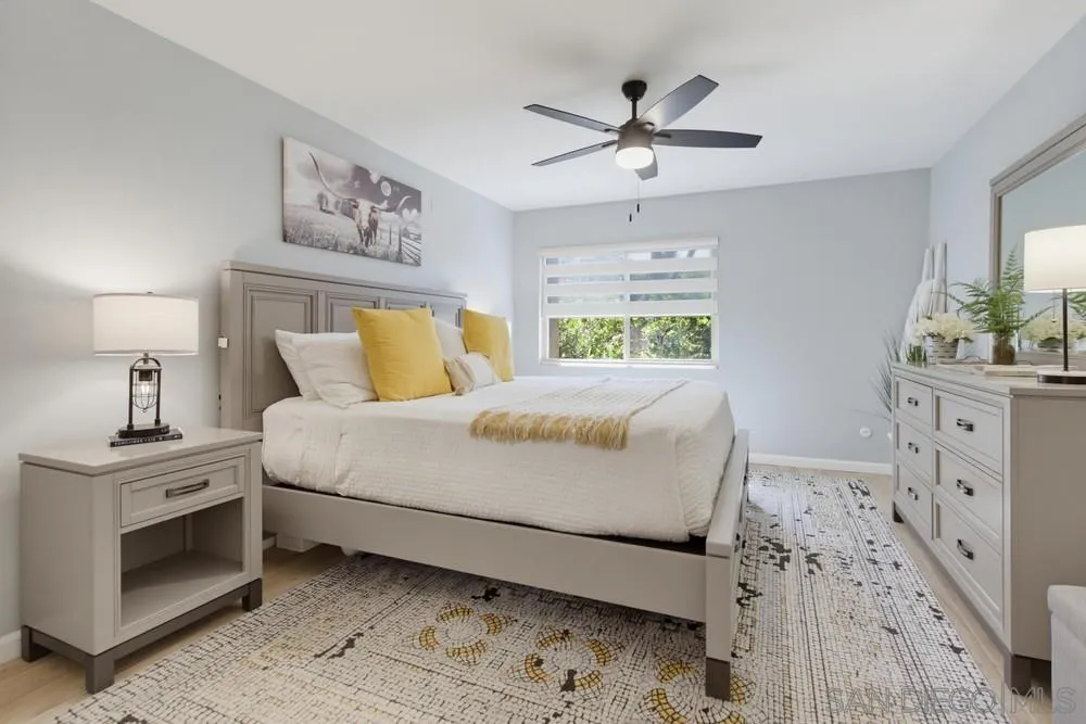 Staged bedroom with a large bed, yellow accent pillows, gray furniture, and a patterned rug.