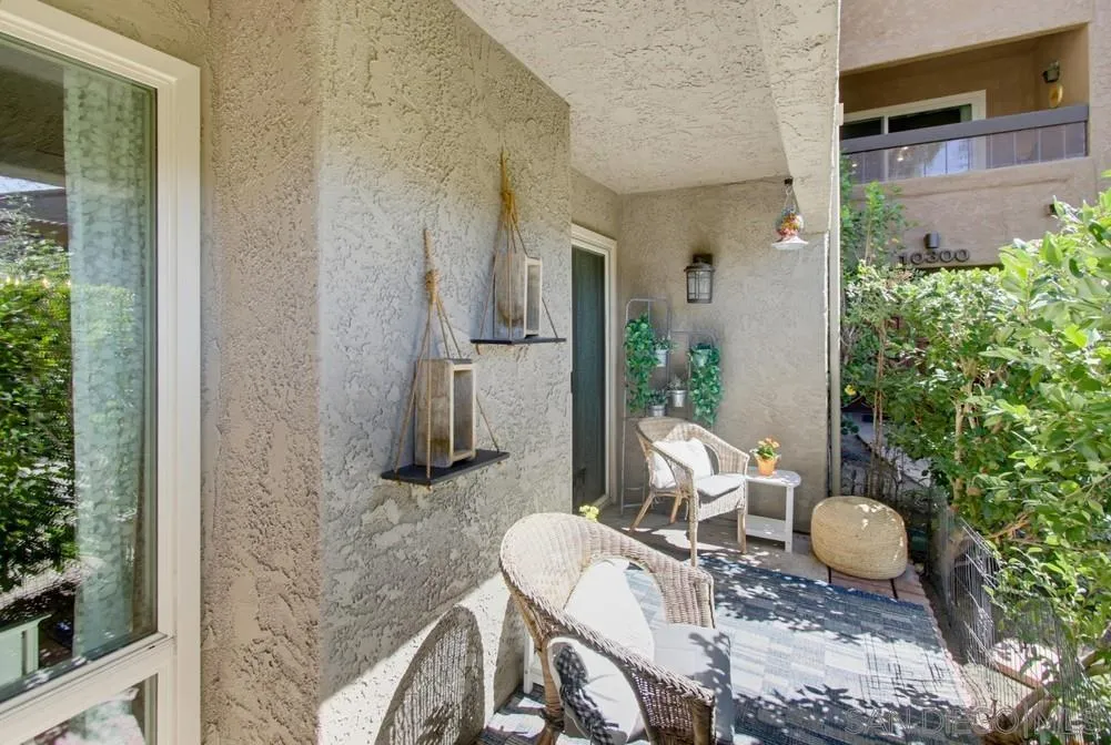 Covered patio with wicker seating, a patterned rug, and greenery beside a stucco wall.
