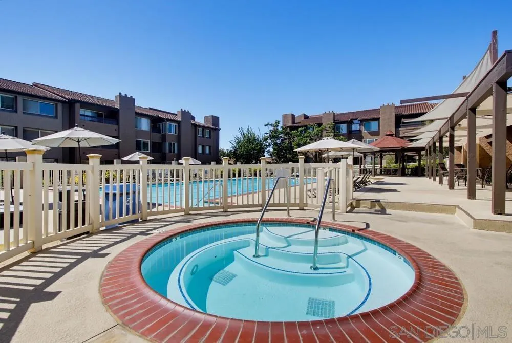 Circular spa beside a fenced community pool, with umbrellas and condominium buildings beyond.