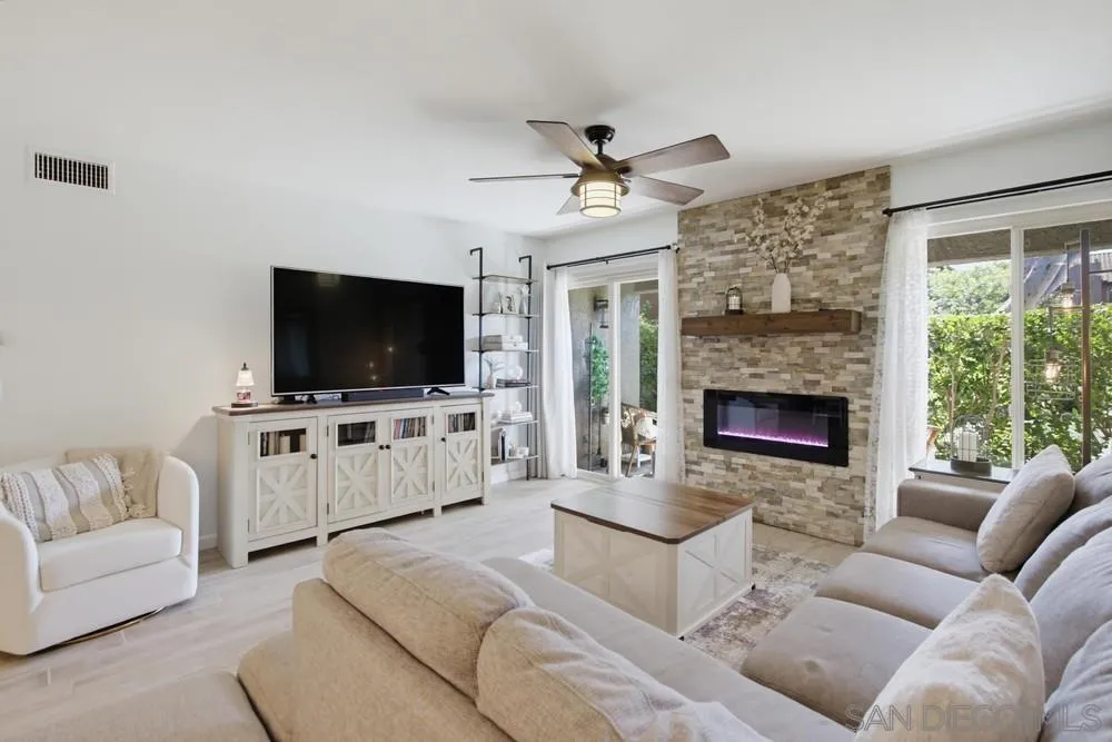 Living room with a stone fireplace, large windows, ceiling fan, and neutral upholstered seating.