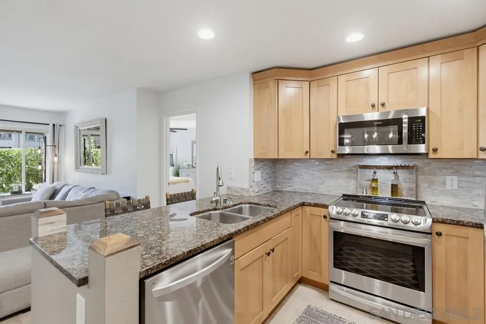 Kitchen with granite counters, light wood cabinets, and stainless steel appliances opening to the living room.