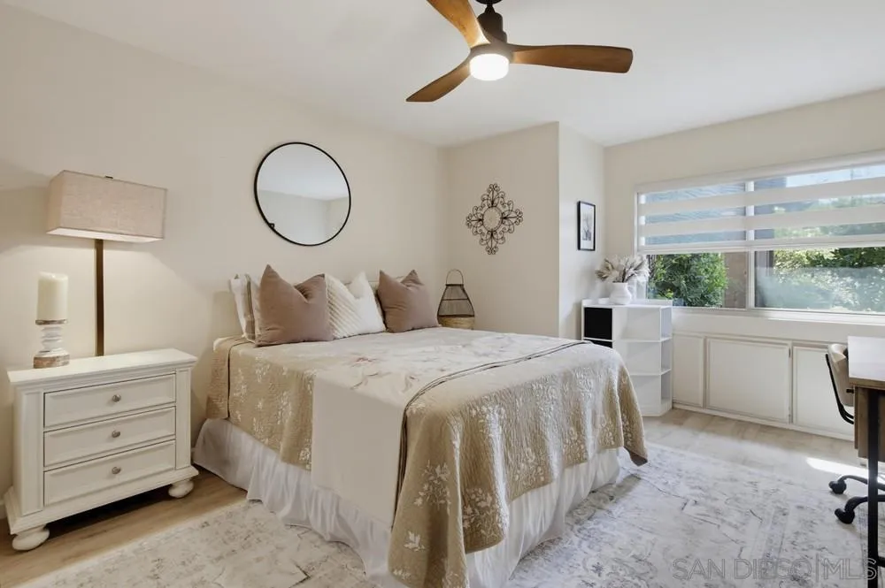 Staged bedroom with a neutral bed, ceiling fan, and wide window overlooking greenery.