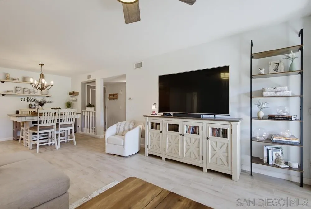 Open living and dining area with pale wood floors, white furniture, built-in shelving, and a large television.