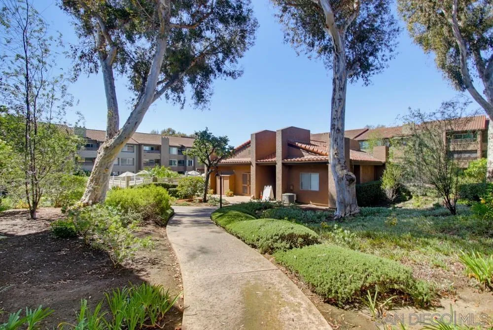 Landscaped walkway leads to a brown stucco condominium building beneath tall trees.